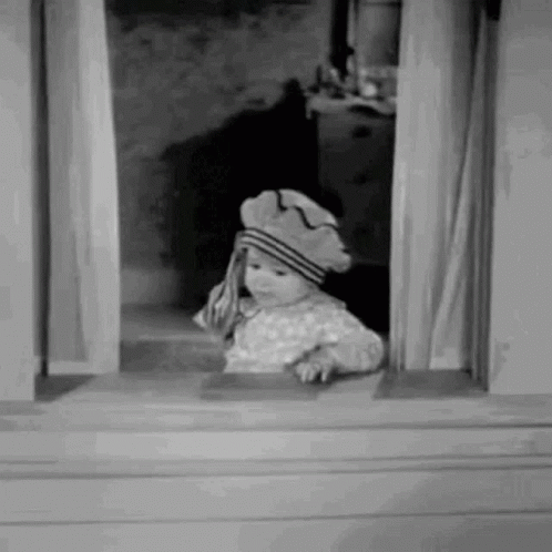 A Little Girl Is Looking Out Of A Window Wearing A Hat . GIF