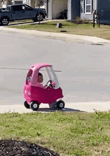 A Little Girl Is Riding A Pink Toy Car Down A Street . GIF