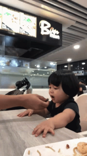 A Little Girl Is Sitting At A Table In Front Of A Sign That Says Bee Mian GIF