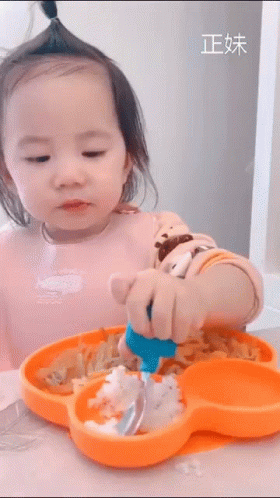 A Little Girl Is Sitting At A Table With A Plate Of Food And A Spoon In Her Hand . GIF