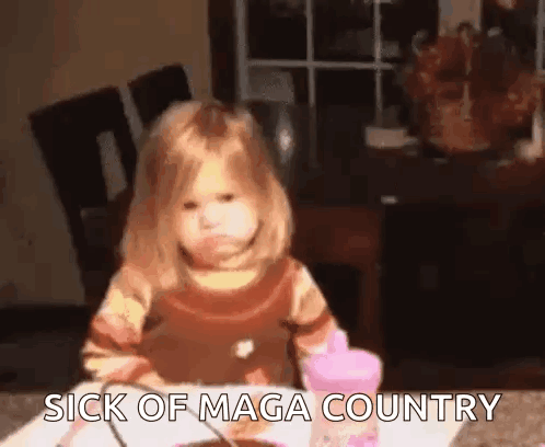 a little girl is sitting at a table with the words `` sick of maga country '' written on the table .