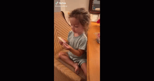 A Little Girl Is Sitting In A Chair Eating A Cookie . GIF