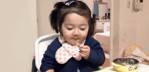A Little Girl Is Sitting In A High Chair Eating A Piece Of Food . GIF