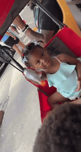 A Little Girl Is Sitting In A Red Cart With A Man Behind Her GIF
