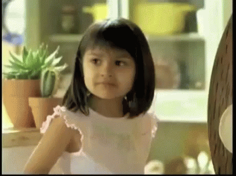 A Little Girl Is Sitting In Front Of A Shelf With Potted Plants In The Background . GIF