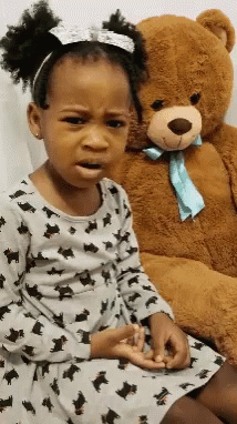 A Little Girl Is Sitting Next To A Teddy Bear And Making A Sad Face GIF