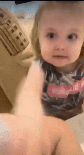 A Little Girl Is Sitting On A Couch Looking At The Camera While A Person Holds Her Hand . GIF