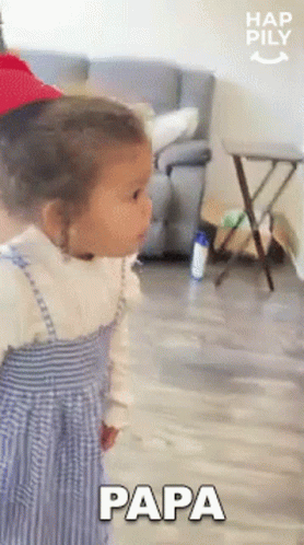 A Little Girl Is Standing In A Living Room Wearing A Red Hat And A Blue Dress And Saying Papa . GIF