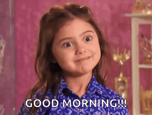 A Little Girl Is Wearing A Blue Shirt And Making A Face And Saying Good Morning . GIF