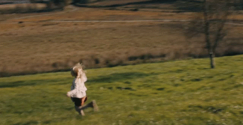 A Little Girl Running Through A Grassy Field With Trees In The Background GIF
