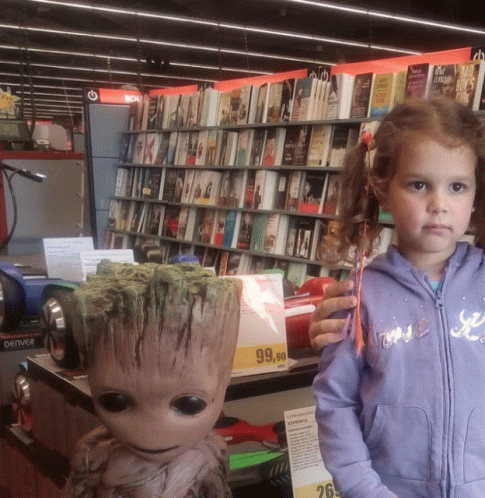 A Little Girl Stands Next To A Statue Of Groot In A Book Store GIF