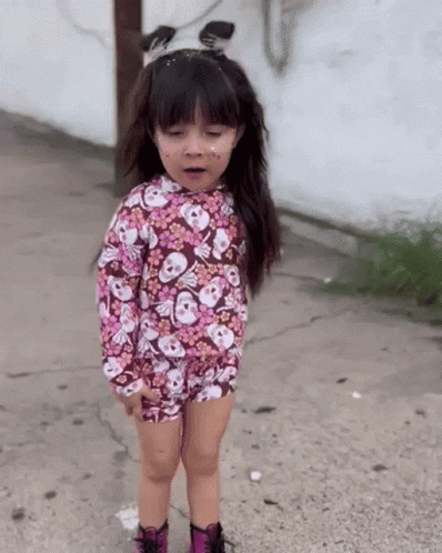 A Little Girl Wearing A Floral Shirt With Skulls On It Is Standing On A Sidewalk GIF