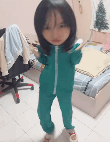 A Little Girl Wearing A Green Jumpsuit Is Standing In A Bedroom GIF