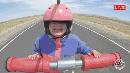 A Little Girl Wearing A Pink Helmet And A Blue Jacket Is Riding A Red Pipe On A Road With A Live Sign Above GIF
