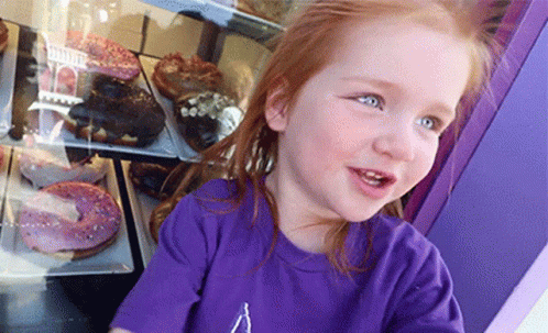 A Little Girl Wearing A Purple Shirt Is Looking At Donuts GIF
