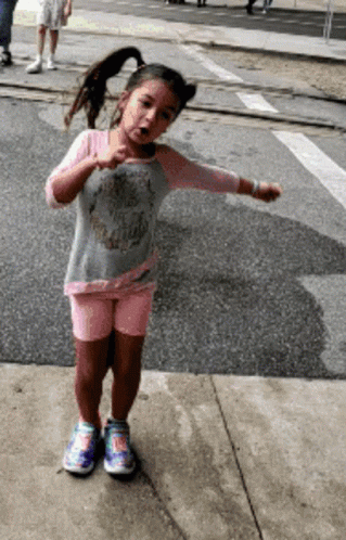 A Little Girl Wearing A Skull Shirt And Shorts Is Standing On The Sidewalk GIF