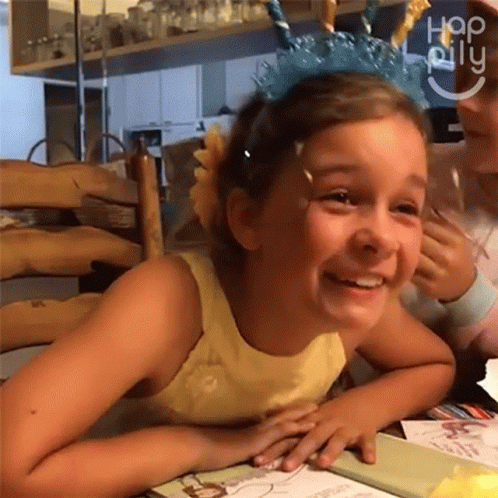 A Little Girl Wearing A Unicorn Headband Smiles While Sitting At A Table . GIF