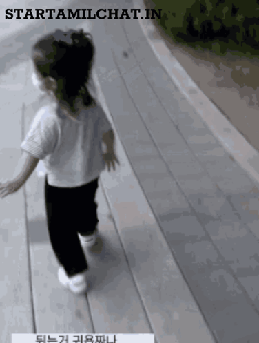 A Little Girl Wearing A White Shirt And Black Pants Is Walking Down A Wooden Walkway GIF