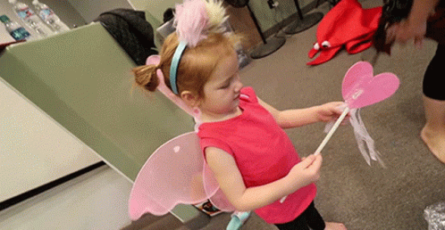 A Little Girl Wearing Fairy Wings And A Headband Holds A Pink Wand GIF
