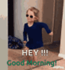 A Little Girl Wearing Sunglasses And A Blue Dress Is Standing In A Doorway And Saying Good Morning . GIF