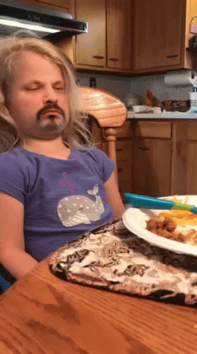 A Little Girl With A Fake Mustache Is Sitting At A Table With A Plate Of Food In Front Of Her GIF