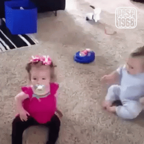 A Little Girl With A Pacifier In Her Mouth Is Sitting Next To A Baby . GIF