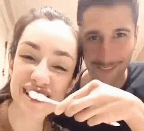 A Man And A Woman Are Brushing Their Teeth Together And Smiling For The Camera . GIF