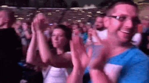 A Man And A Woman Are Clapping Their Hands In A Crowd . GIF