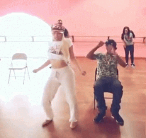 A Man And A Woman Are Dancing In A Dance Studio While A Man Sits In A Chair . GIF