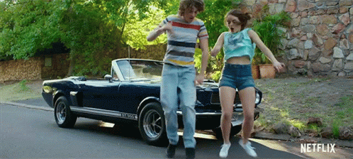 A Man And A Woman Are Dancing In Front Of A Blue Mustang Convertible . GIF