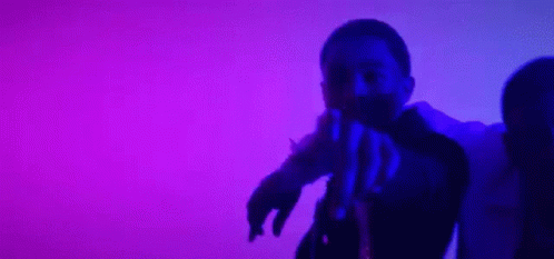 A Man And A Woman Are Dancing In Front Of A Purple Background . The Man Is Pointing At The Camera . GIF