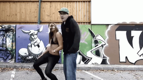 A Man And A Woman Are Dancing In Front Of A Wall With Graffiti Including The Letter N. GIF