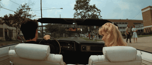 A Man And A Woman Are Driving Down A Street In A Car With A School In The Background GIF
