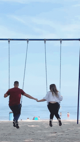 A Man And A Woman Are Holding Hands While Sitting On Swings GIF