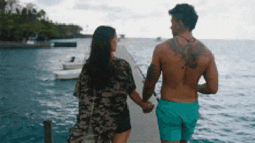 A Man And A Woman Are Holding Hands While Walking On A Dock Overlooking The Ocean . GIF