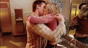 A Man And A Woman Are Hugging And Kissing In A Bathroom In Front Of A Mirror . GIF