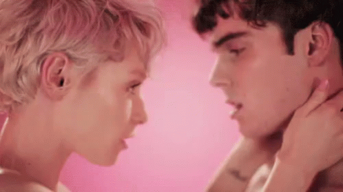 A Man And A Woman Are Kissing In Front Of A Pink Background . The Woman Is Touching The Man 'S Neck . GIF