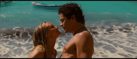 A Man And A Woman Are Kissing On A Beach With A Boat In The Background . GIF