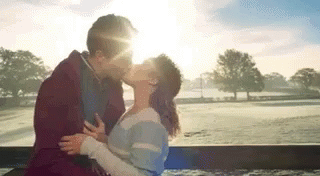 A Man And A Woman Are Kissing While Sitting On A Bench In Front Of A Field . GIF