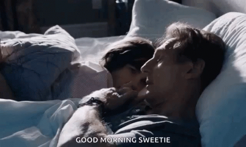 A Man And A Woman Are Laying In Bed And The Man Is Kissing The Woman On The Cheek . GIF