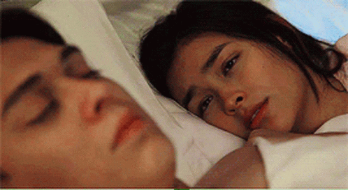 A Man And A Woman Are Laying In Bed Together And The Woman Is Looking At The Man . GIF