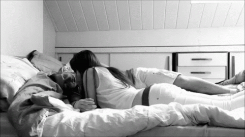 A Man And A Woman Are Laying On A Bed With White Sheets GIF
