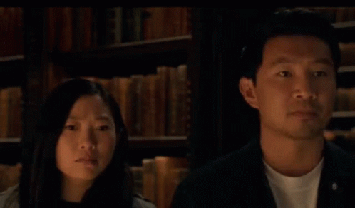 A Man And A Woman Are Looking At Each Other In Front Of A Bookshelf With The Words Or Written In Blue . GIF
