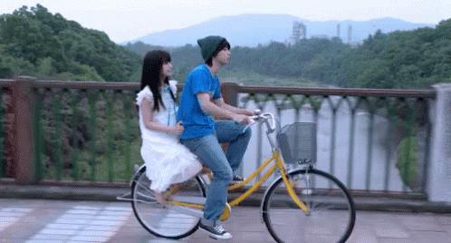 A Man And A Woman Are Riding A Bicycle On A Bridge Over A Body Of Water . GIF