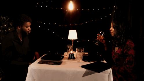 A Man And A Woman Are Sitting At A Table Drinking Wine And Talking GIF