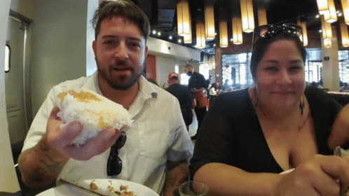 A Man And A Woman Are Sitting At A Table Eating Food GIF