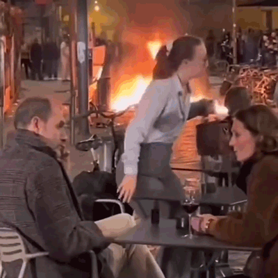 A Man And A Woman Are Sitting At A Table In Front Of A Burning Building . GIF