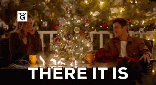 A Man And A Woman Are Sitting At A Table In Front Of A Christmas Tree With The Words There It Is Above Them . GIF
