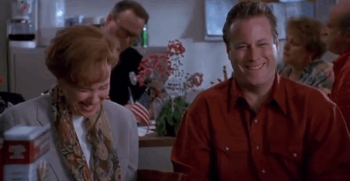 A Man And A Woman Are Sitting At A Table Laughing And Smiling . GIF