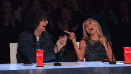 A Man And A Woman Are Sitting At A Table Laughing . The Woman Is Pointing At The Man . GIF